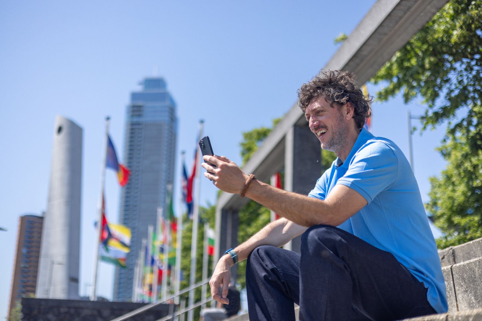 Medewerker glimlacht naar zijn smartphone bij de vlaggen aan de Boompjes in Rotterdam.